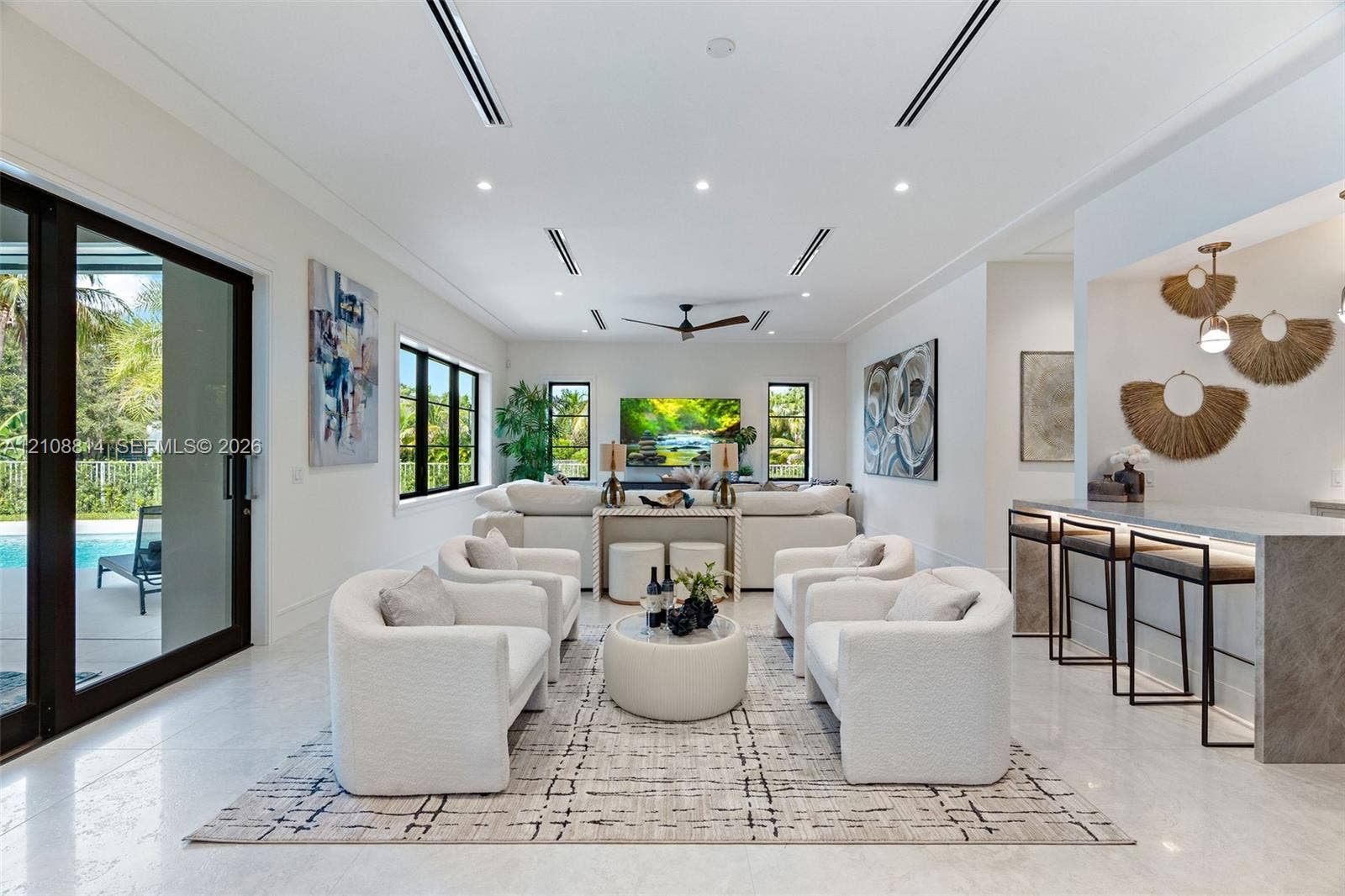 Open-concept living area featuring large-format stone flooring, recessed linear lighting, black-framed windows, integrated wet bar with stone countertop, and multi-panel sliding glass doors