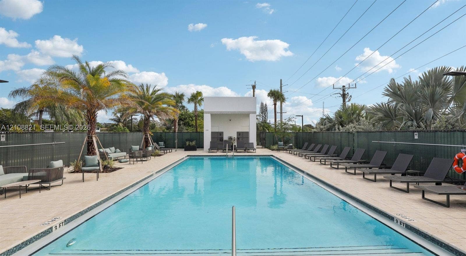 Rectangular swimming pool featuring a tiled deck, perimeter fencing, palm tree landscaping, and a central cabana structure