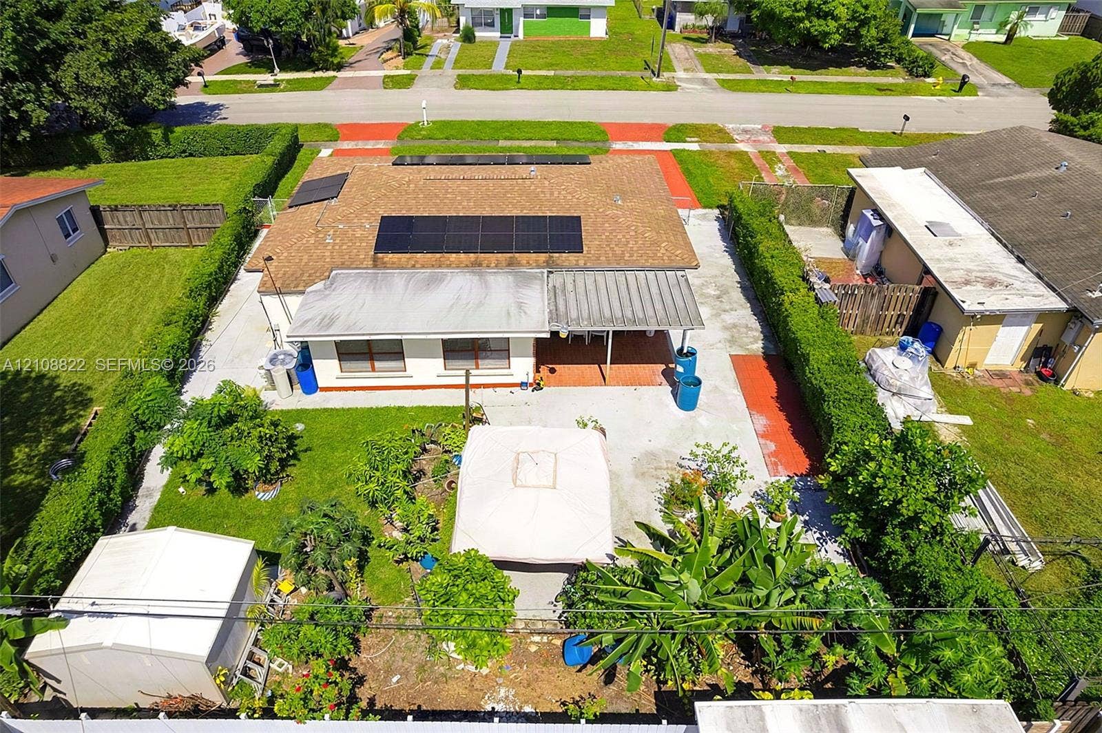 6838 SW 22nd Court, Miramar, FL 33023 Exterior featuring a shingle roof equipped with solar panels, a covered patio, a concrete rear yard, and mature perimeter hedging
