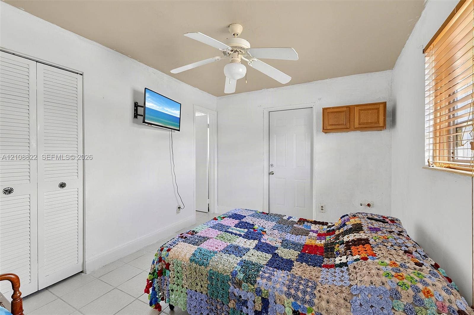 6838 SW 22nd Court, Miramar, FL 33023 Bedroom featuring white louvered-panel closet doors, neutral-toned tile flooring, ceiling fan with integrated lighting, and wall-mounted wooden cabinetry