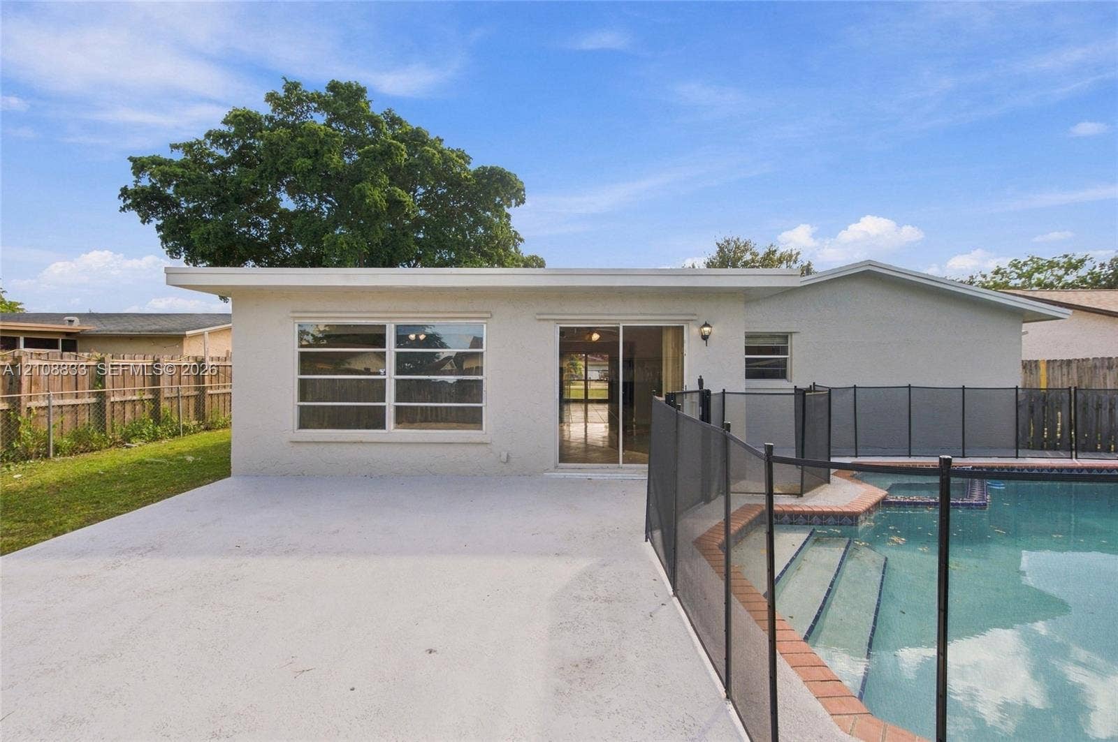 9611 Johnson Street, Pembroke Pines, FL 33024 Rear exterior featuring a concrete patio, a swimming pool with tiered steps, an integrated safety fence, and a wood-slat perimeter fence