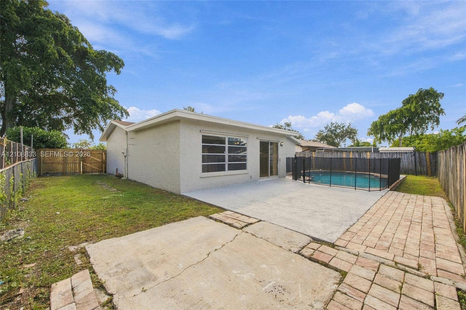 9611 Johnson Street, Pembroke Pines, FL 33024 Backyard featuring an in-ground pool with mesh safety fencing, a concrete patio, brick paver accents, and wood perimeter fencing