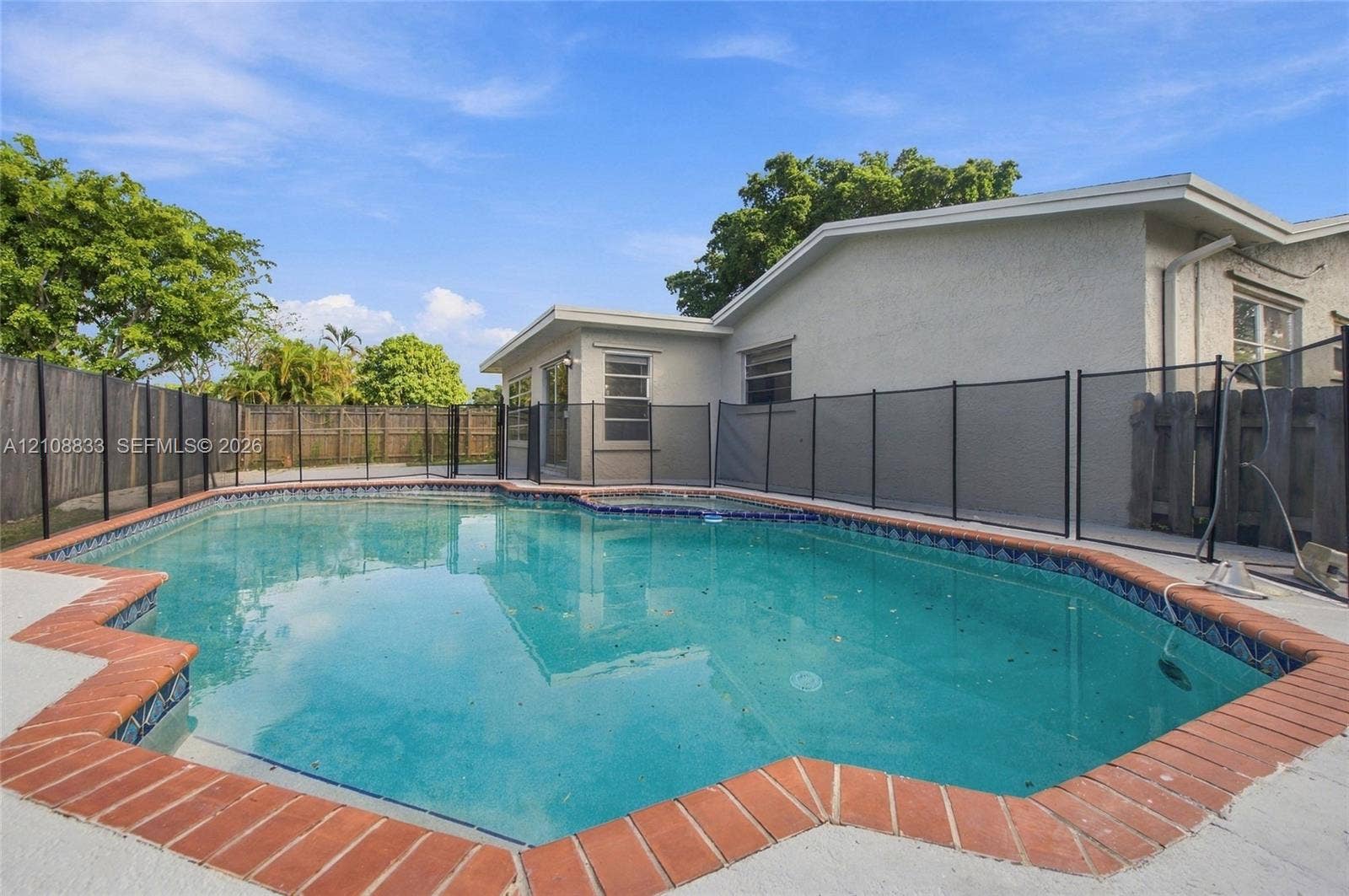 9611 Johnson Street, Pembroke Pines, FL 33024 In-ground swimming pool featuring a brick paver surround, decorative tile border, and a black mesh safety fence enclosed by wood privacy fencing