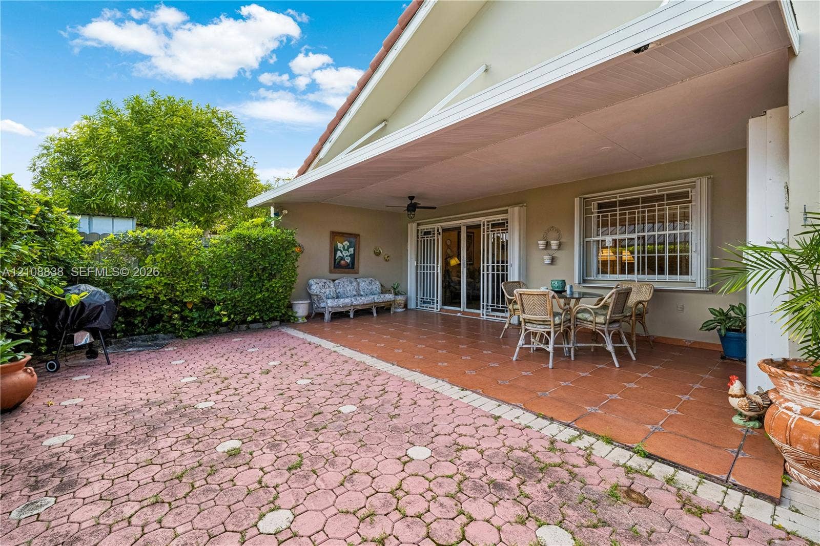Covered patio featuring ceramic tile flooring, a ceiling fan, and a patterned paver outdoor area enclosed by mature hedge landscaping