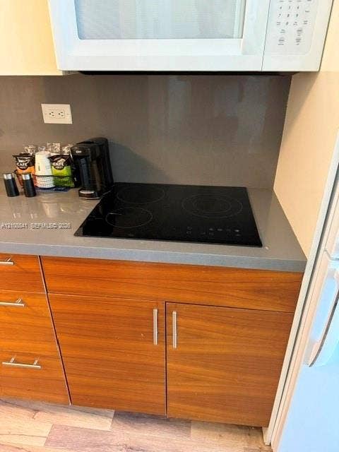 Kitchen area featuring a smooth-surface electric cooktop, solid-surface gray countertops, matching full-height backsplash, and wood-finish cabinetry with brushed metal hardware