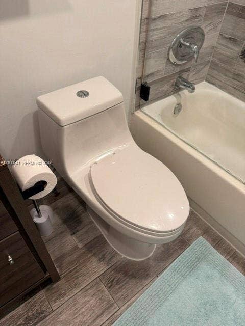 One-piece elongated toilet featuring a dual-flush button, wood-finish tile flooring, and a bathtub surround with grey-toned textured tile and chrome fixtures