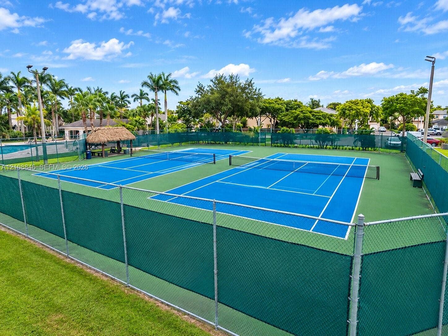 11827 SW 99th Lane, Miami, FL 33186 Dual tennis courts featuring high-tensile chain-link fencing, a thatched-roof pavilion, and integrated court lighting