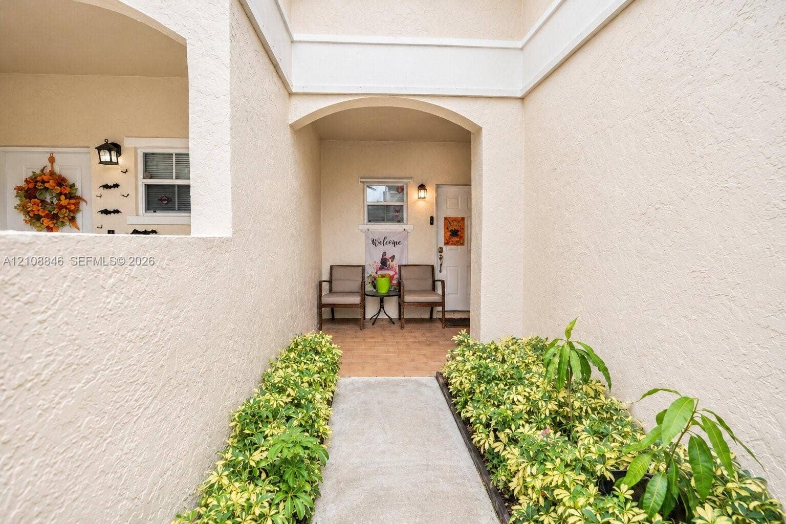 11827 SW 99th Lane, Miami, FL 33186 Stucco exterior entryway featuring a concrete walkway, arched architectural detailing, wall-mounted light fixtures, and manicured perimeter landscaping