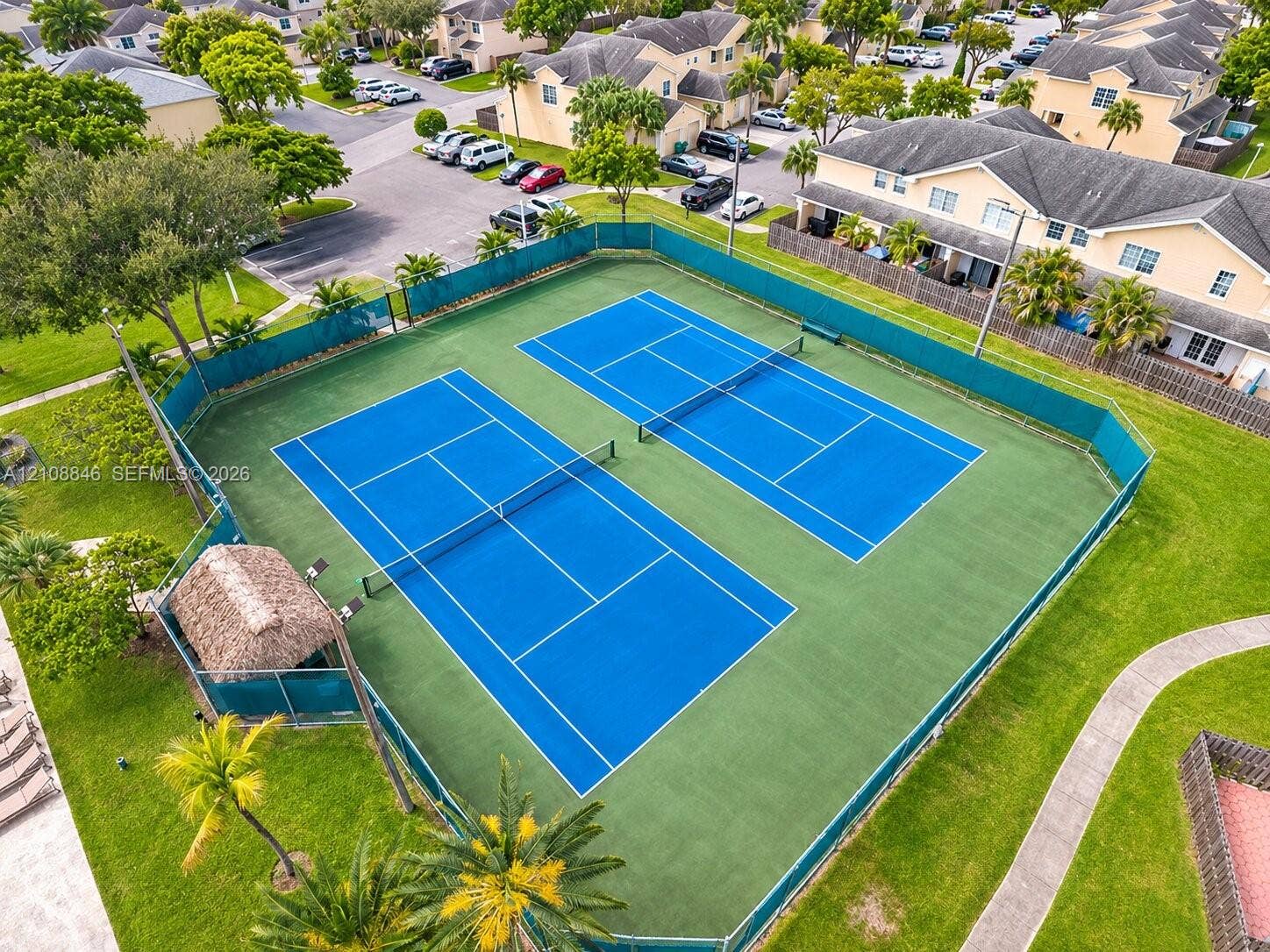 11827 SW 99th Lane, Miami, FL 33186 Dual tennis courts featuring hard-surface playing areas, perimeter fencing, and an integrated thatched-roof structure