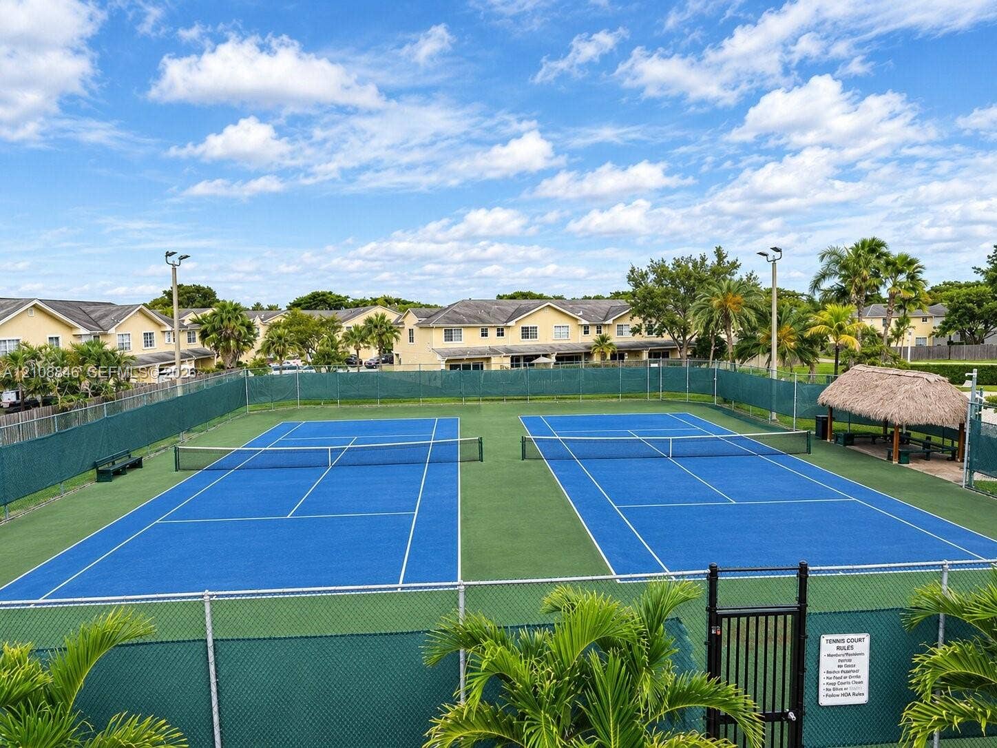 11827 SW 99th Lane, Miami, FL 33186 Dual tennis courts featuring specialized surface markings, perimeter chain-link fencing, stadium-style lighting, and a thatched-roof pavilion