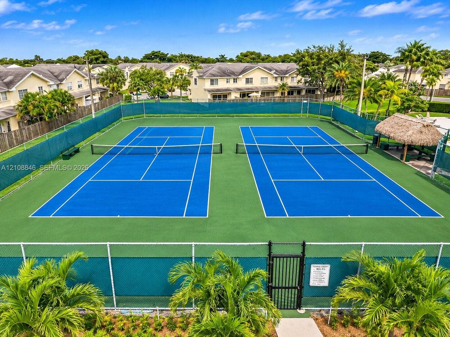 11827 SW 99th Lane, Miami, FL 33186 Dual tennis courts featuring professional surfacing, perimeter chain-link fencing, and a thatch-roofed cabana