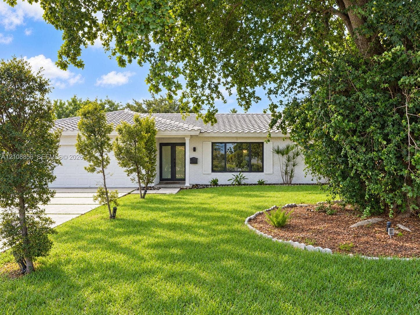 Single-story residence featuring a white painted brick exterior, tile roof, expansive front lawn, landscaped mulch garden beds with stone border, and a concrete driveway