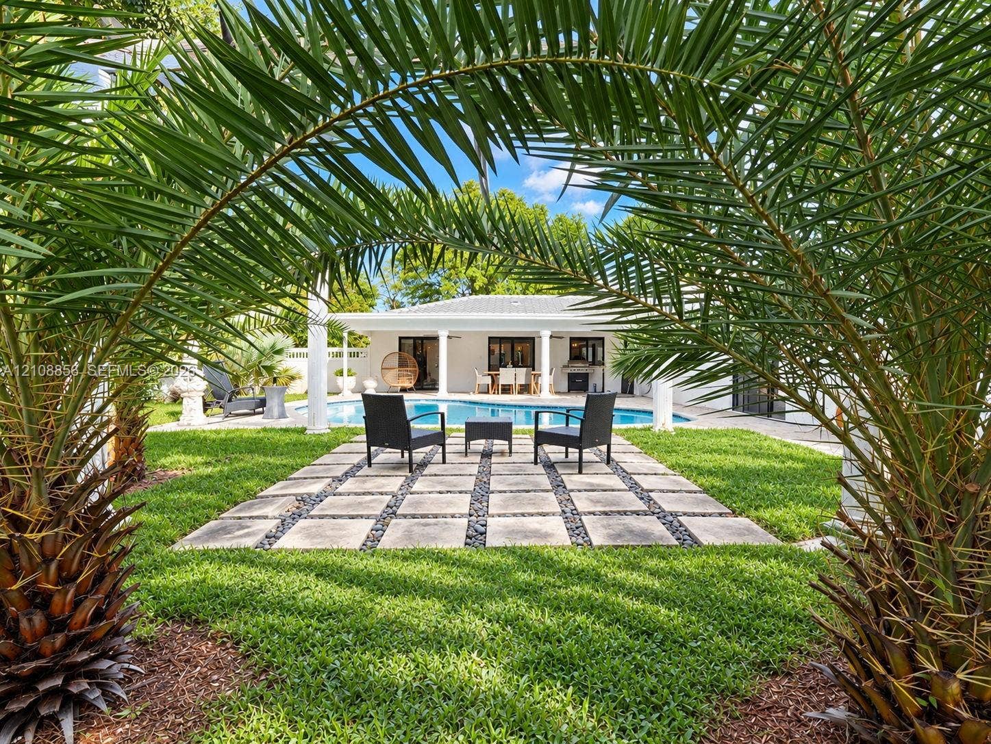 Backyard featuring a paved stone patio with pebble accents, a swimming pool, mature palm trees, and a covered terrace supported by columns