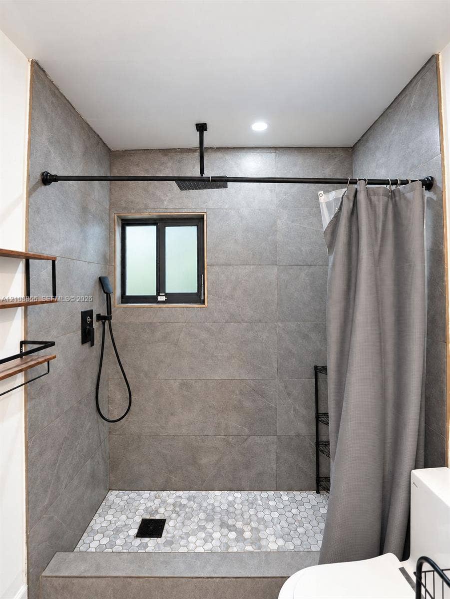 Walk-in shower featuring large-format grey tile surround, hexagonal floor tiling, a ceiling-mounted rainfall showerhead, and matte black plumbing fixtures