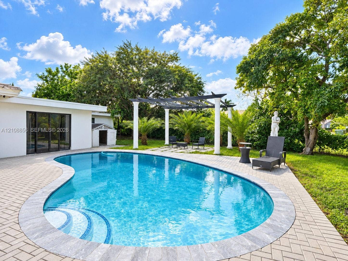 Custom-shaped swimming pool with tiered entry steps, herringbone-patterned stone paver patio, and pergola supported by white columns