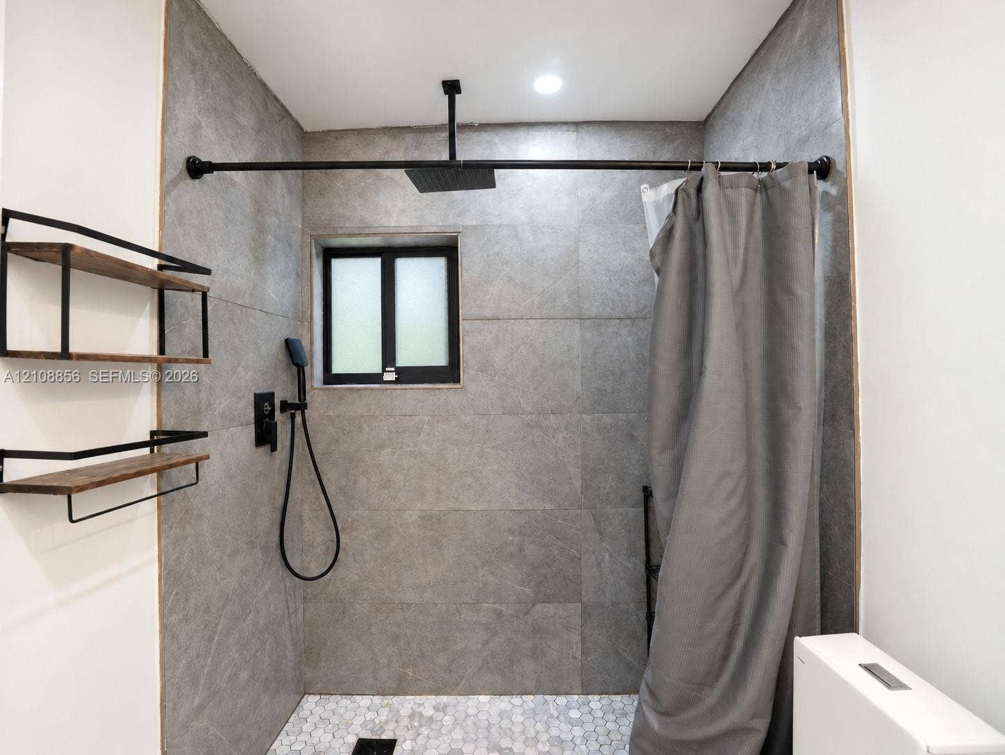 Shower area featuring large-format grey tile walls, a white hexagonal tile floor, matte black rainfall showerhead with handheld sprayer, and built-in wood-finish shelving with black metal framing