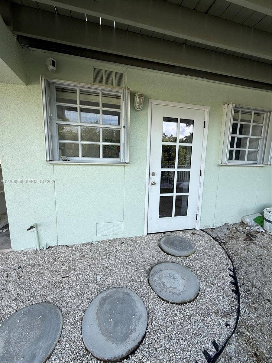 Exterior entry featuring a glass-paneled door, dual grid-style windows, pebble ground cover, and circular concrete pavers