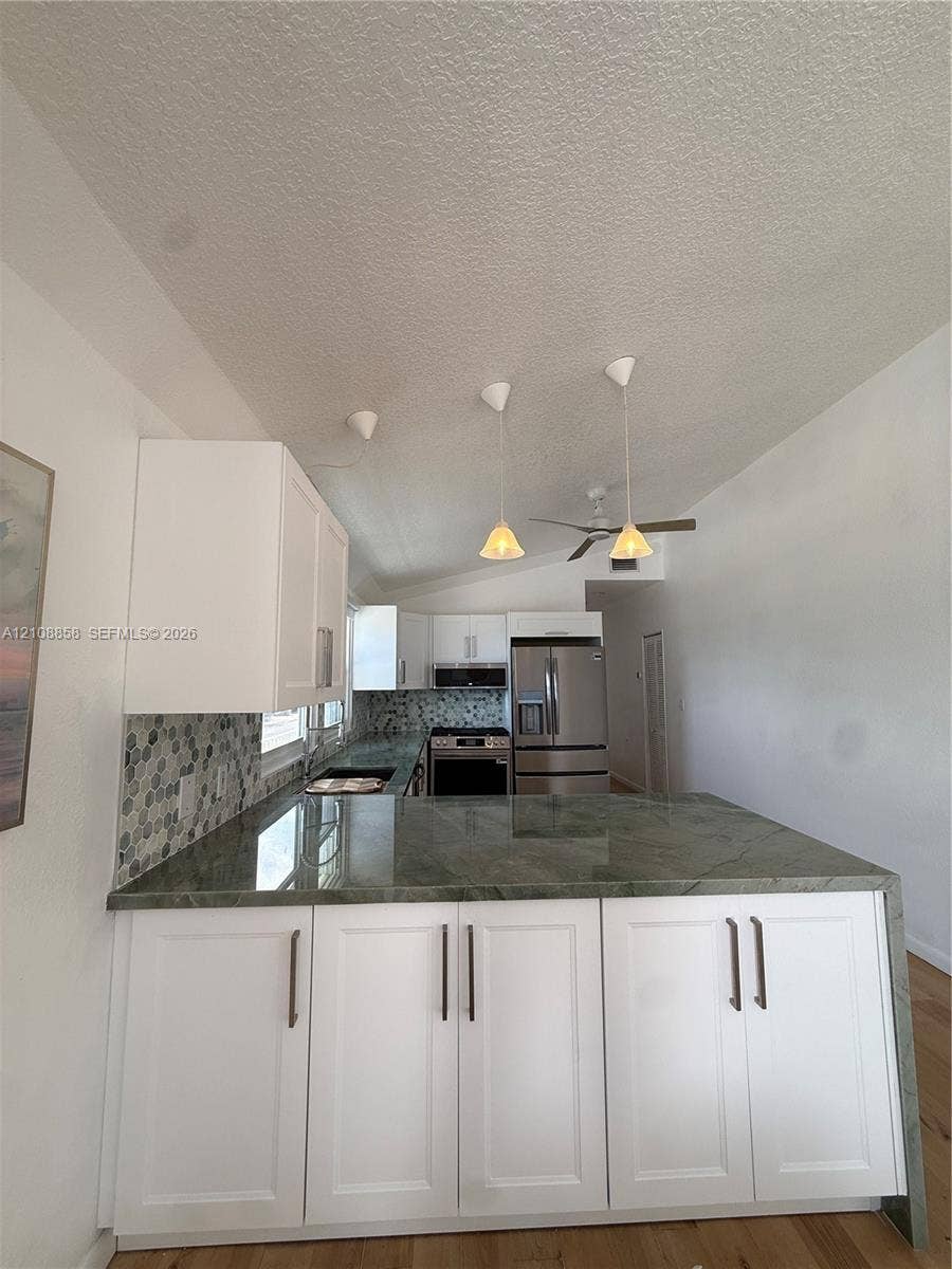 Kitchen featuring white shaker-style cabinetry, solid-surface countertops with waterfall edge, geometric tile backsplash, stainless steel appliances, and pendant lighting