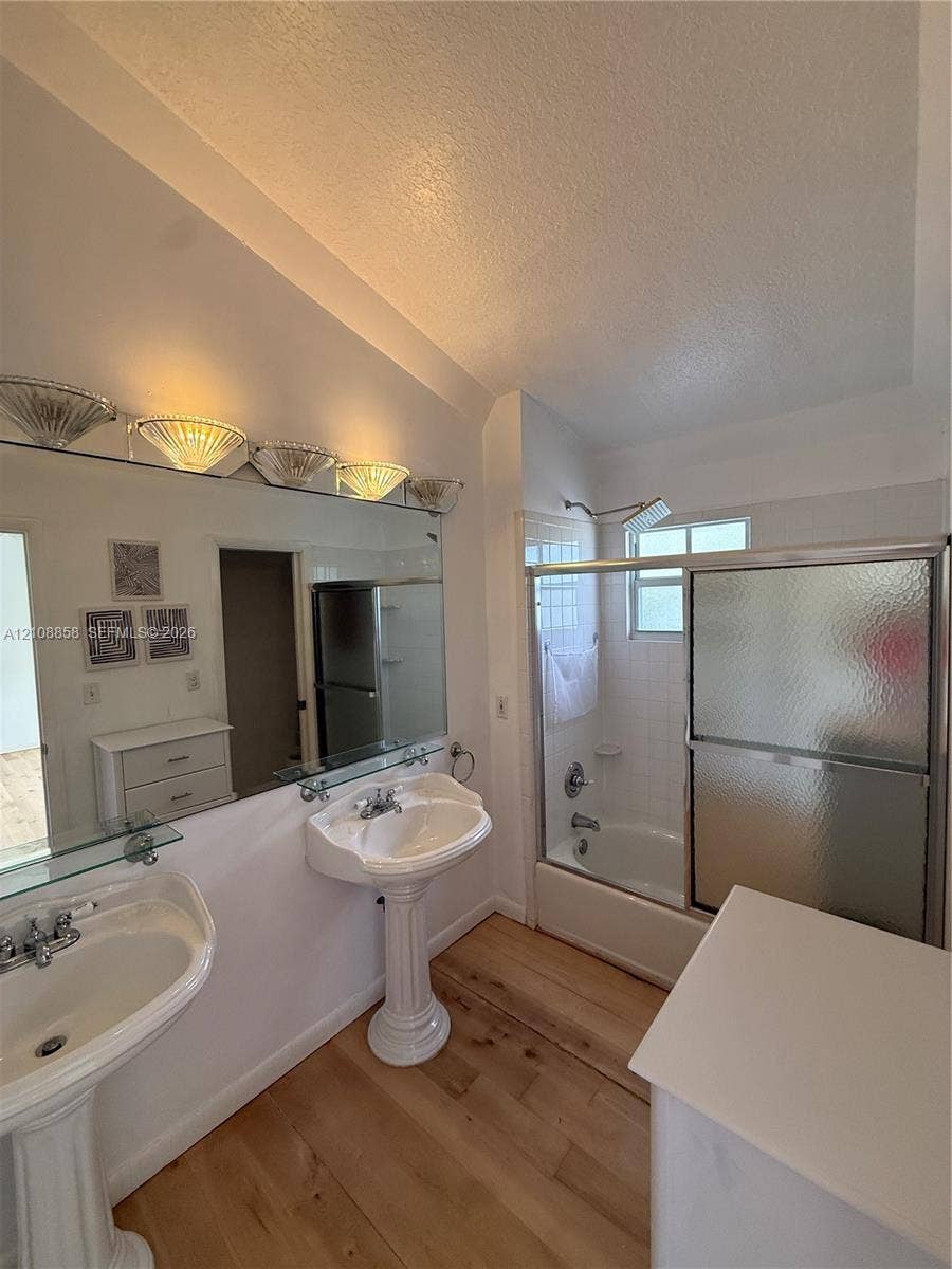 Dual porcelain pedestal sinks, wood-finish flooring, full-width wall mirror, and a tub-shower combination with sliding glass doors