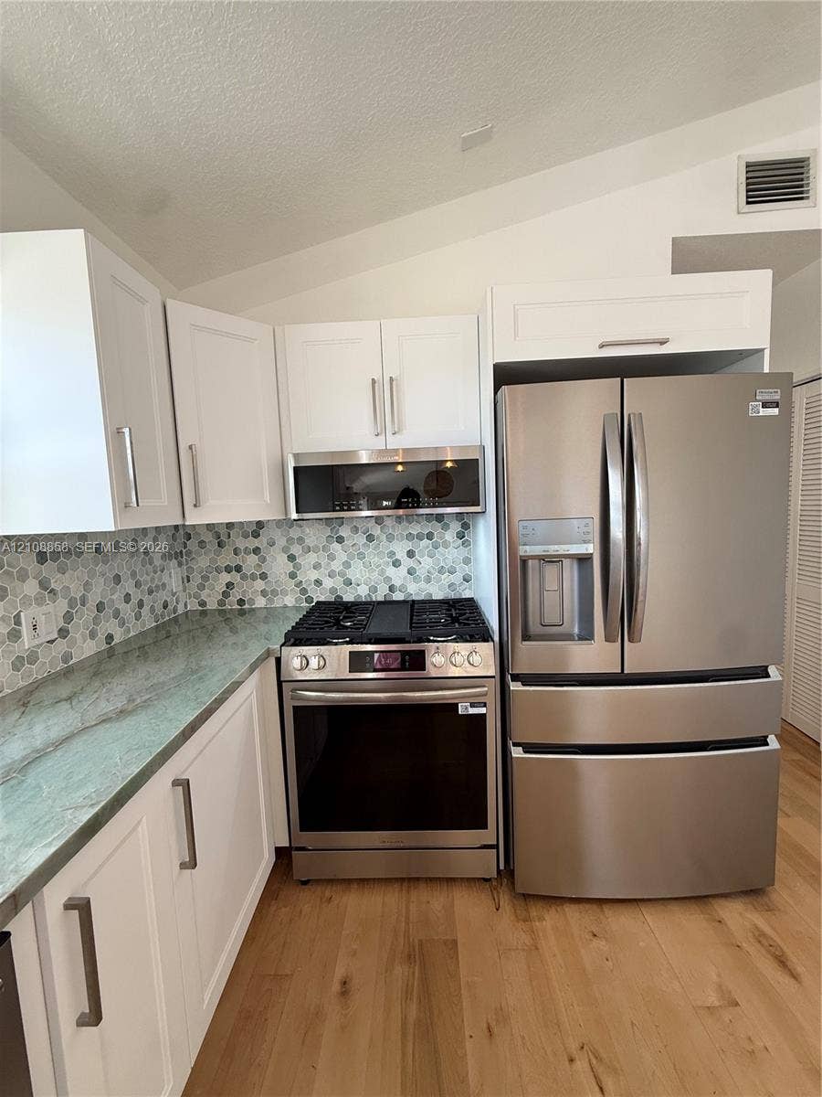 Kitchen featuring white shaker-style cabinetry, natural stone countertops, hexagonal tile backsplash, stainless steel appliances, and wood-finish flooring