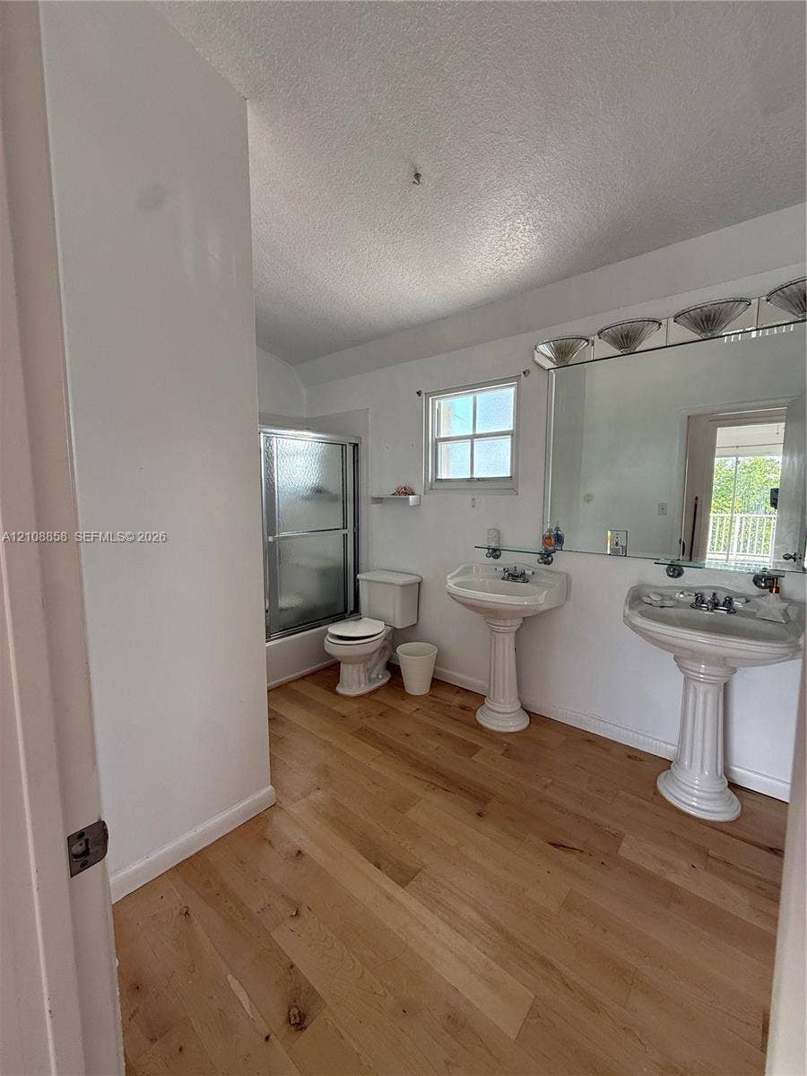 Bathroom featuring wood-finish flooring, dual pedestal sinks, a large wall-mounted mirror with integrated lighting, and a glass-enclosed shower