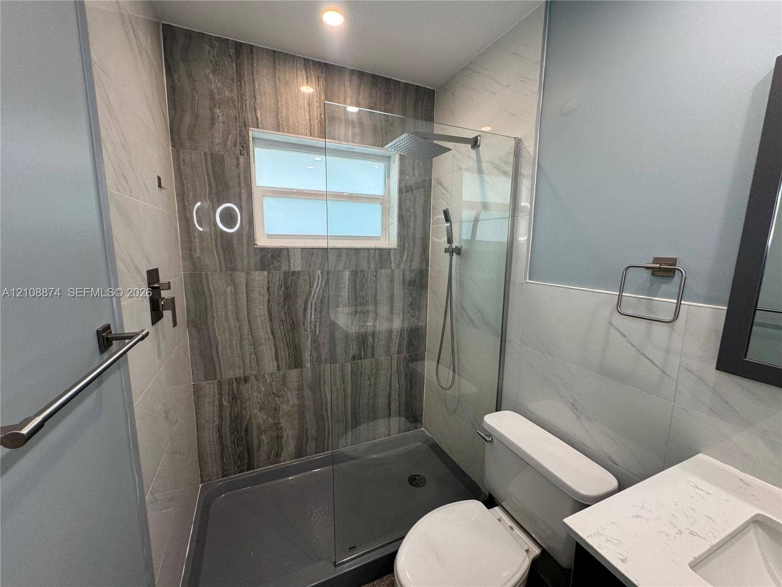 Walk-in shower with a glass panel, dark wood-finish tile surround, ceiling-mounted rainfall showerhead, and a quartz countertop vanity