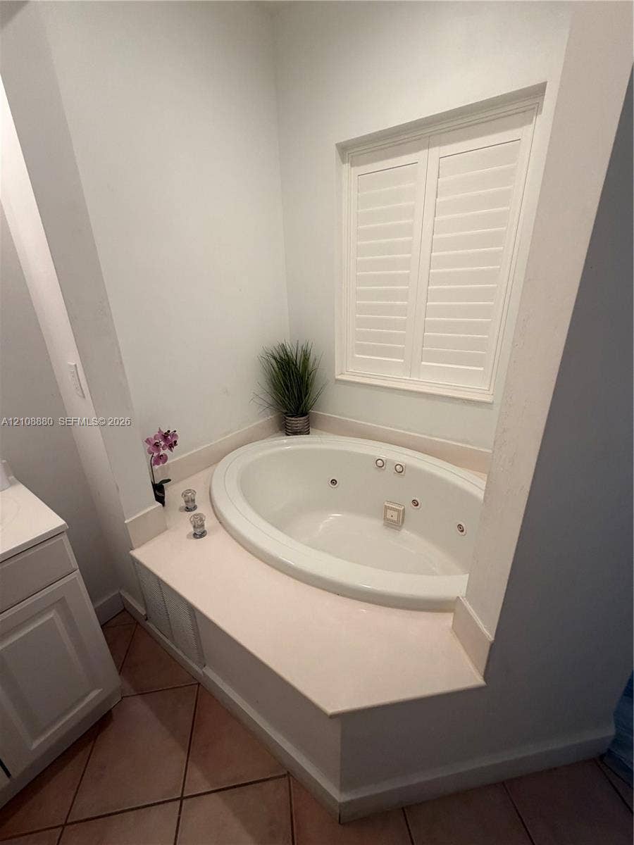 Recessed soaking tub featuring a tiled deck surround and interior jets, accompanied by plantation shutters and ceramic tile flooring