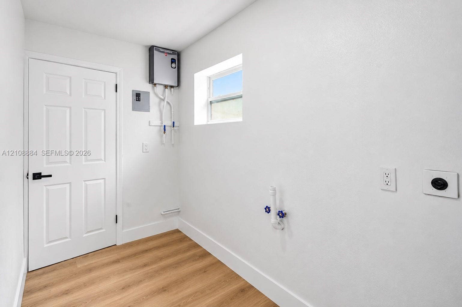 Utility space featuring wood-finish flooring, a wall-mounted tankless water heater, white six-panel door with matte black hardware, and an egress window