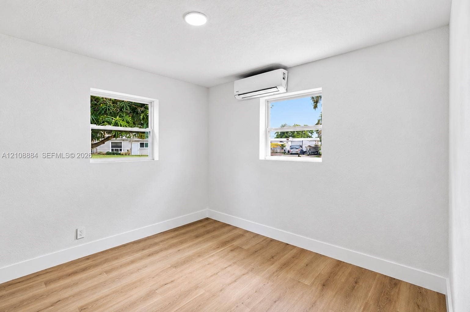 Neutral-toned interior featuring wood-finish flooring, two large windows, a ceiling-mounted light fixture, and a wall-mounted ductless mini-split climate control system