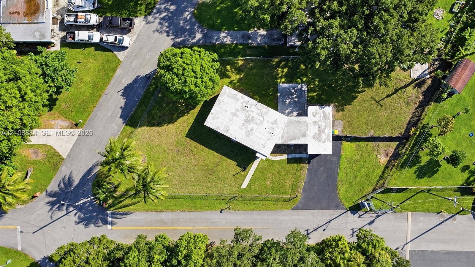 Aerial view of a rectangular property featuring a paved asphalt driveway, expansive green lawn, mature trees, and a flat-roof residence