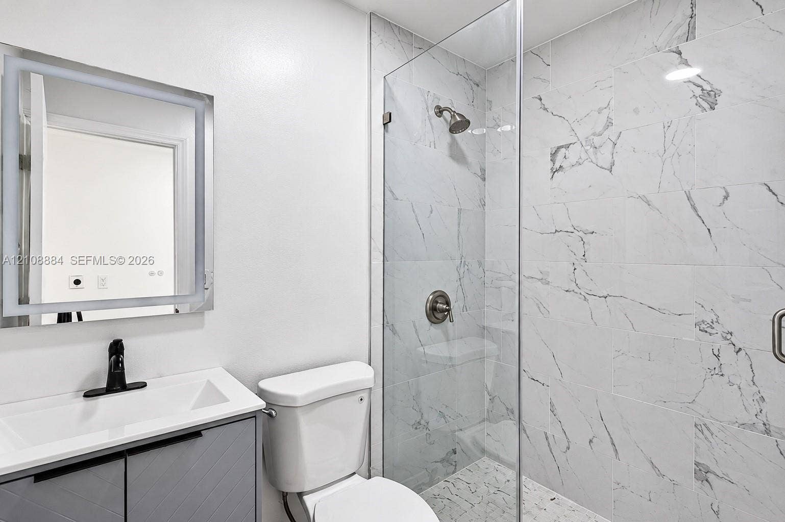 Bathroom featuring a gray vanity with a white countertop, wall-mounted LED-backlit mirror, and walk-in shower with floor-to-ceiling tile surround and glass enclosure