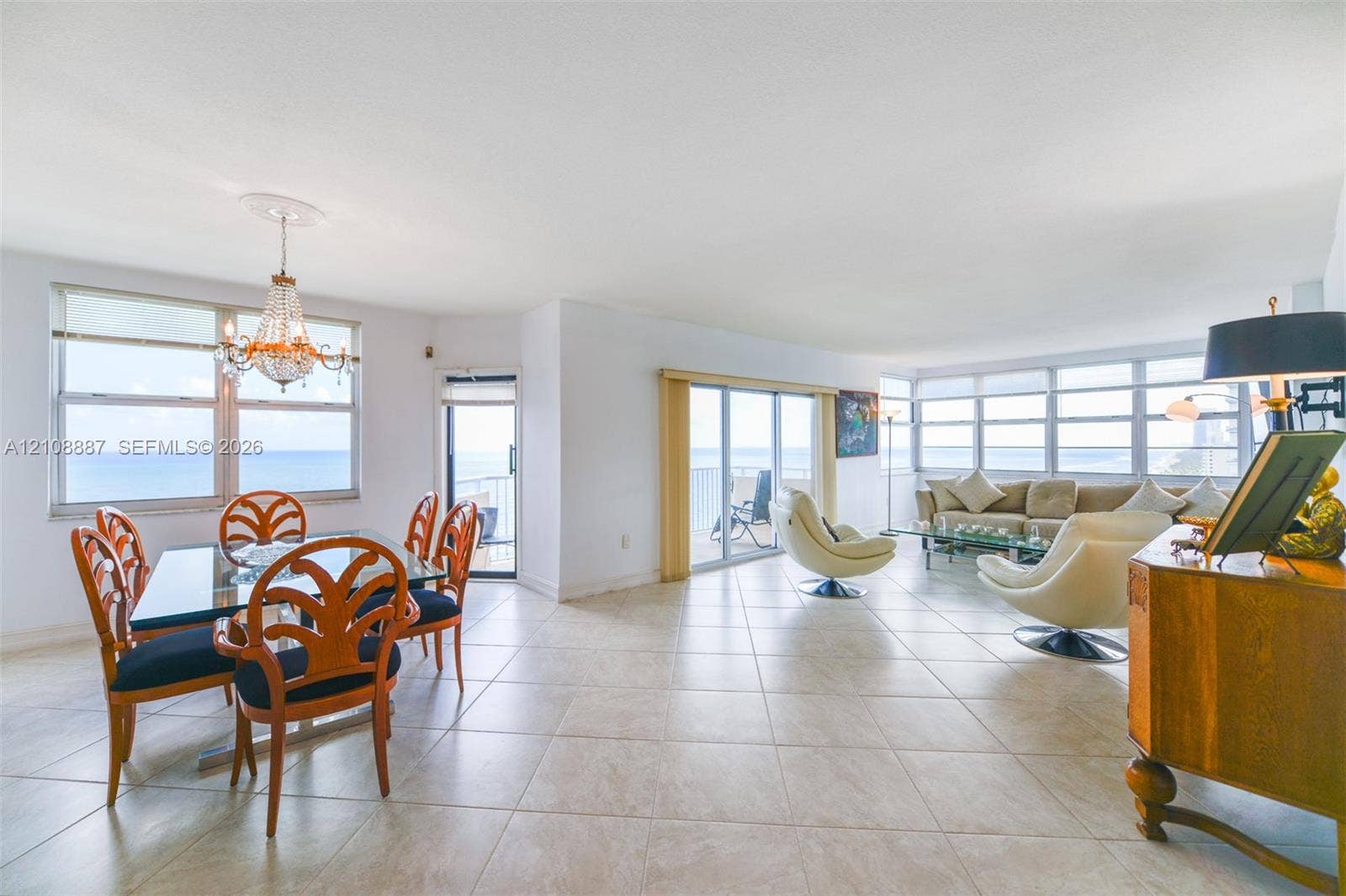 Open-concept main living area featuring light-toned tile flooring, oversized windows, multi-pane sliding glass doors, and a decorative crystal chandelier