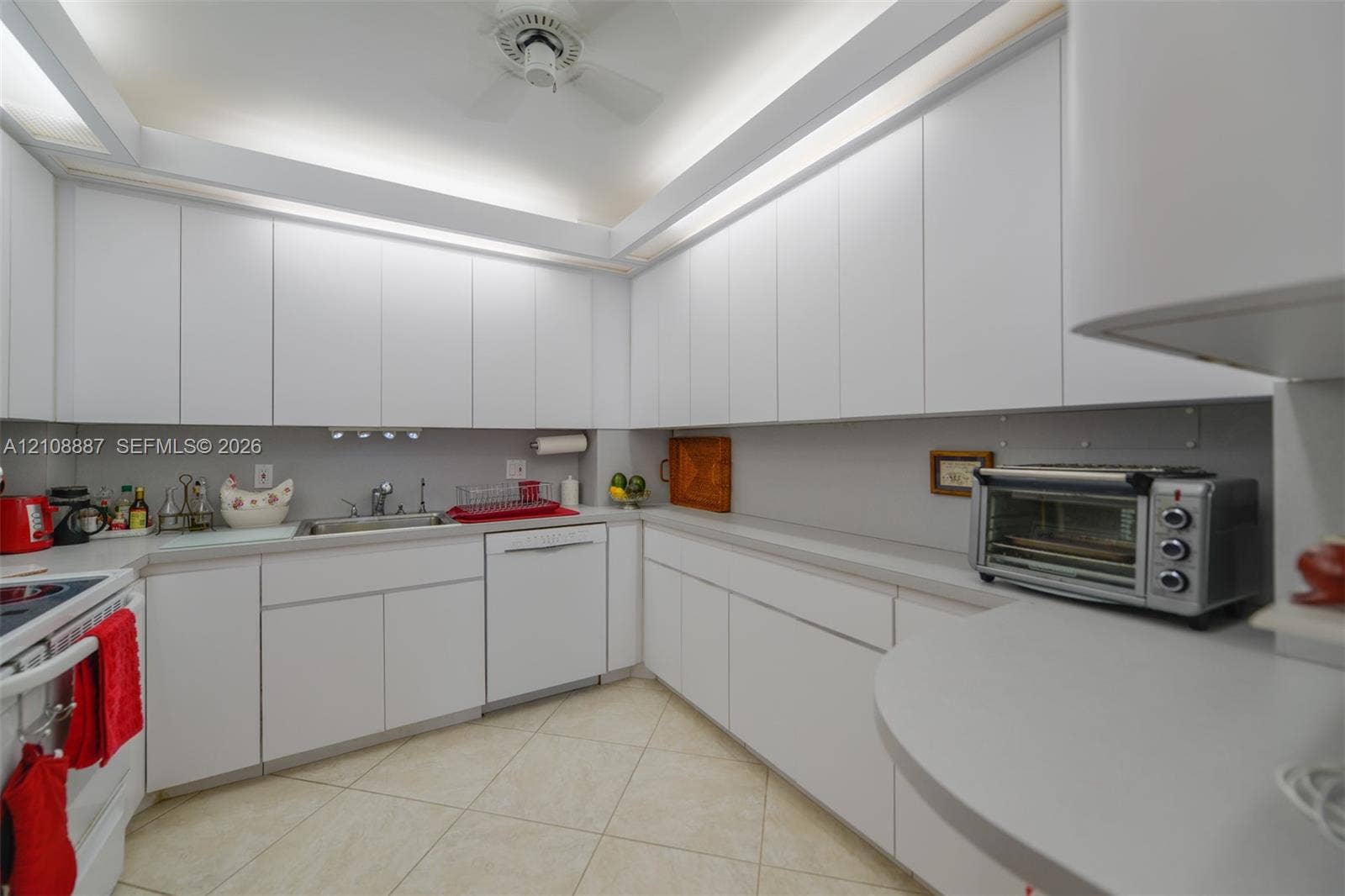 Kitchen featuring floor-to-ceiling white cabinetry, solid-surface countertops, integrated under-cabinet lighting, stainless steel sink with metal faucet, and tile flooring