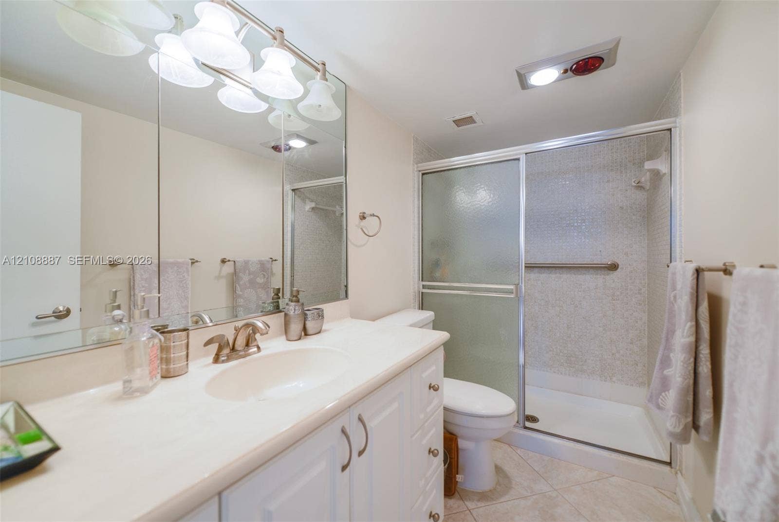 White vanity cabinetry featuring a light-toned countertop with integrated sink, full-width mirror, five-bulb integrated vanity lighting, and walk-in shower with sliding glass doors