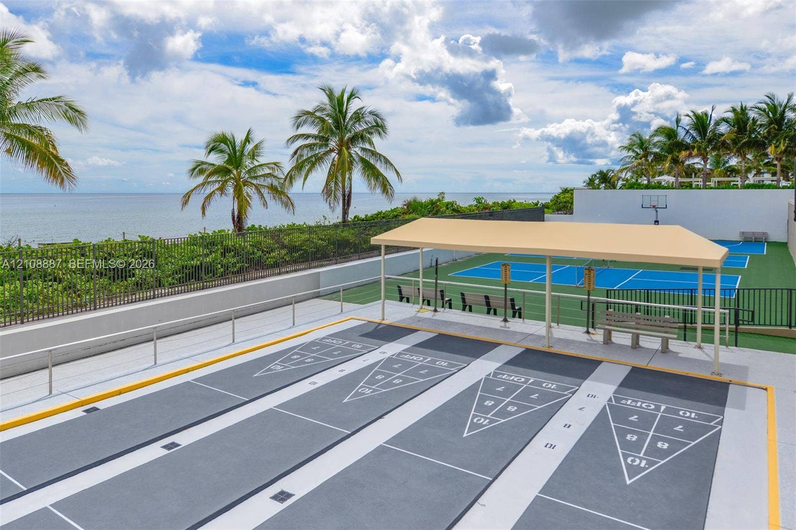 Shuffleboard courts with a shaded pavilion and a hard-surface sports court featuring a basketball hoop, set against an oceanfront backdrop with palm trees