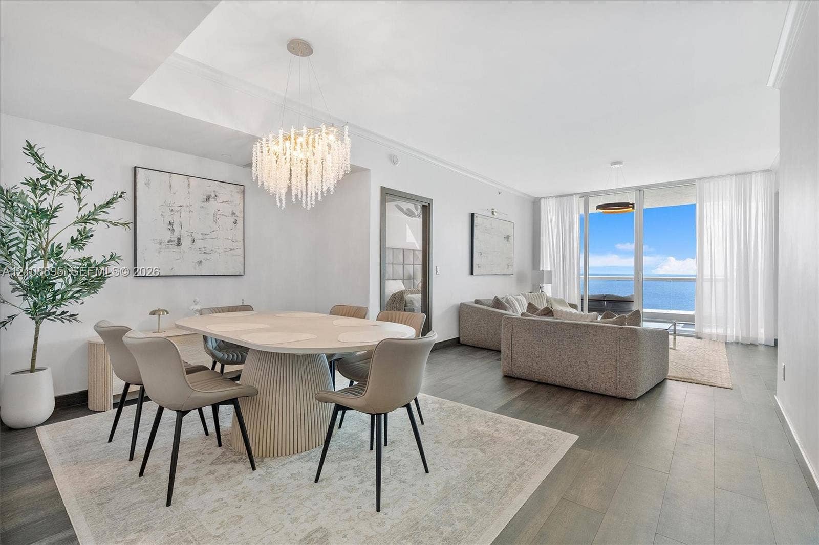 Open-concept interior featuring wood-finish flooring, a tiered crystal chandelier, tray ceiling, and floor-to-ceiling glass sliding doors opening to a balcony