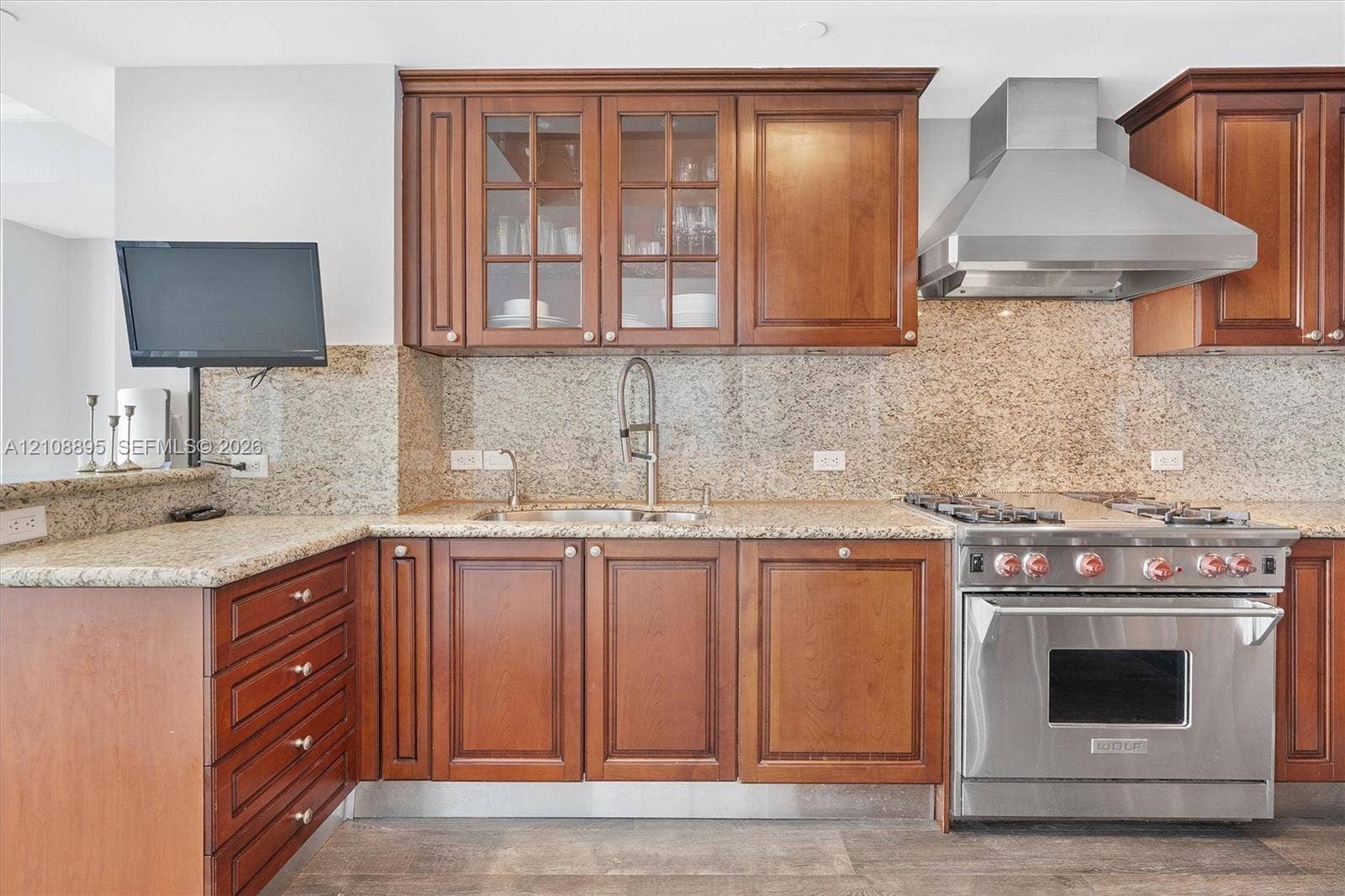 Kitchen featuring custom wood cabinetry, stone countertops with matching backsplash, stainless steel gas range, professional-grade range hood, and glass-front upper cabinet doors