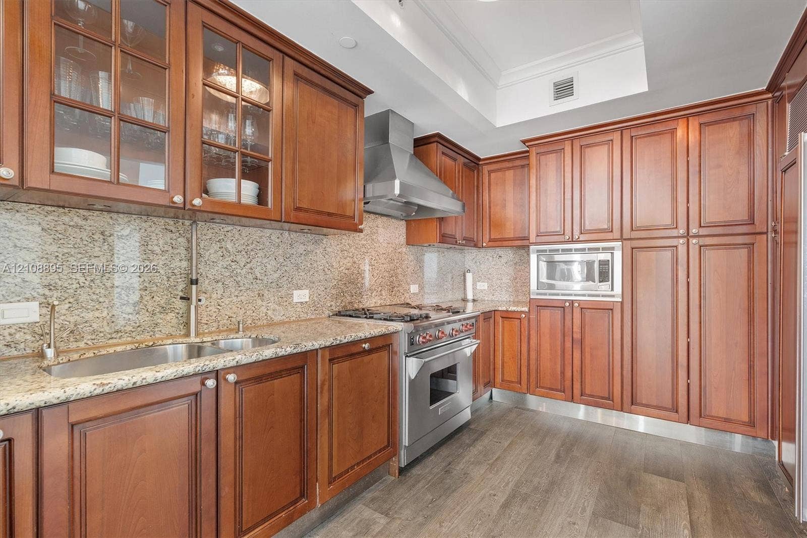 Kitchen featuring custom wood cabinetry, natural stone countertops, a stainless steel gas range with a matching range hood, and wood-finish flooring