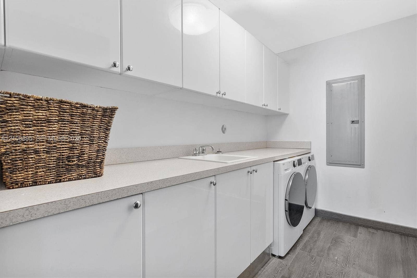 Laundry area featuring high-gloss white upper and lower cabinetry, stone-composite countertop with an integrated sink, metal faucet hardware, and wood-finish flooring