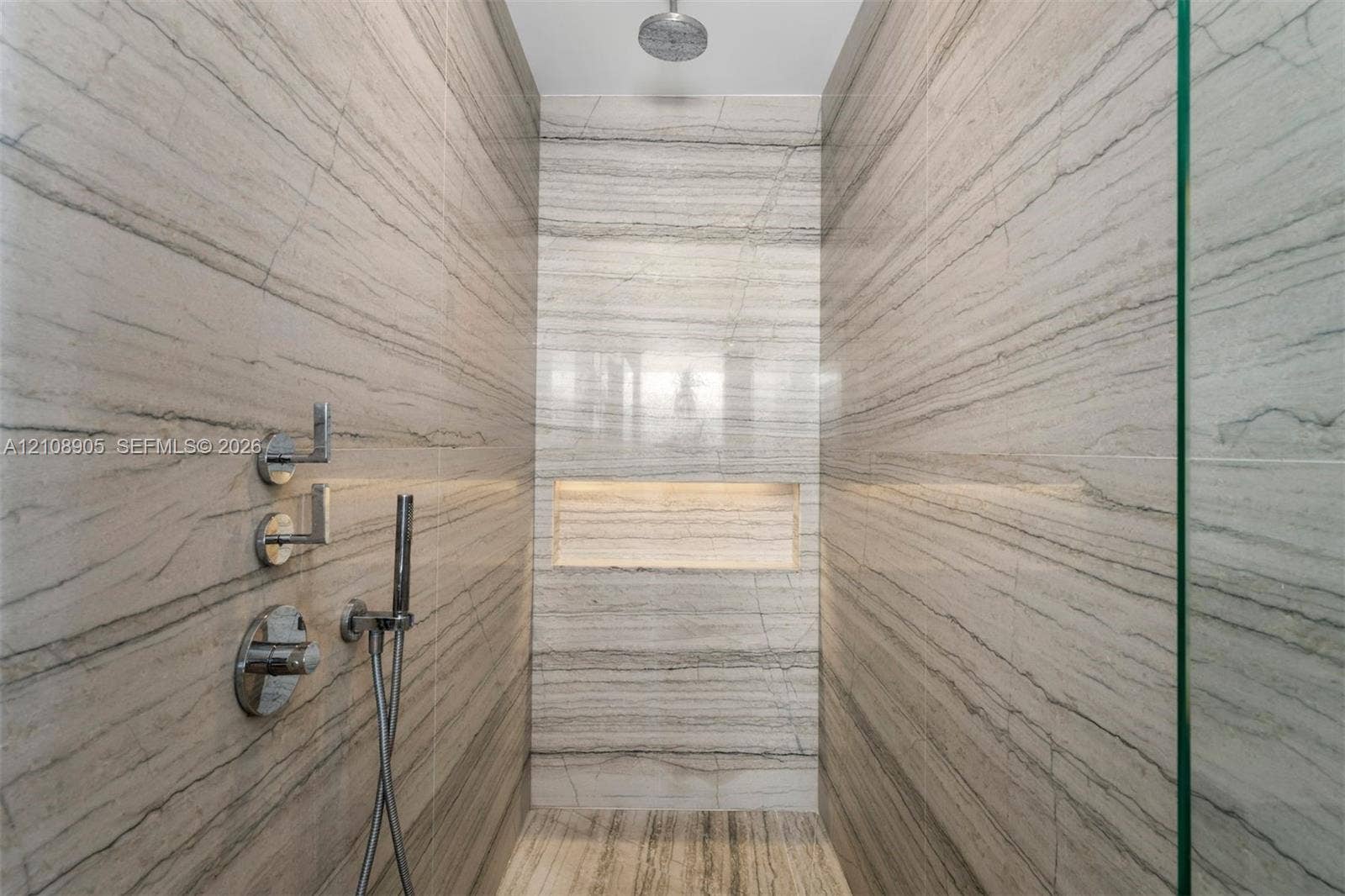 Walk-in shower featuring floor-to-ceiling stone-tiled walls, a built-in recessed niche, chrome wall-mounted fixtures, a handheld sprayer, and a ceiling-mounted rainfall showerhead