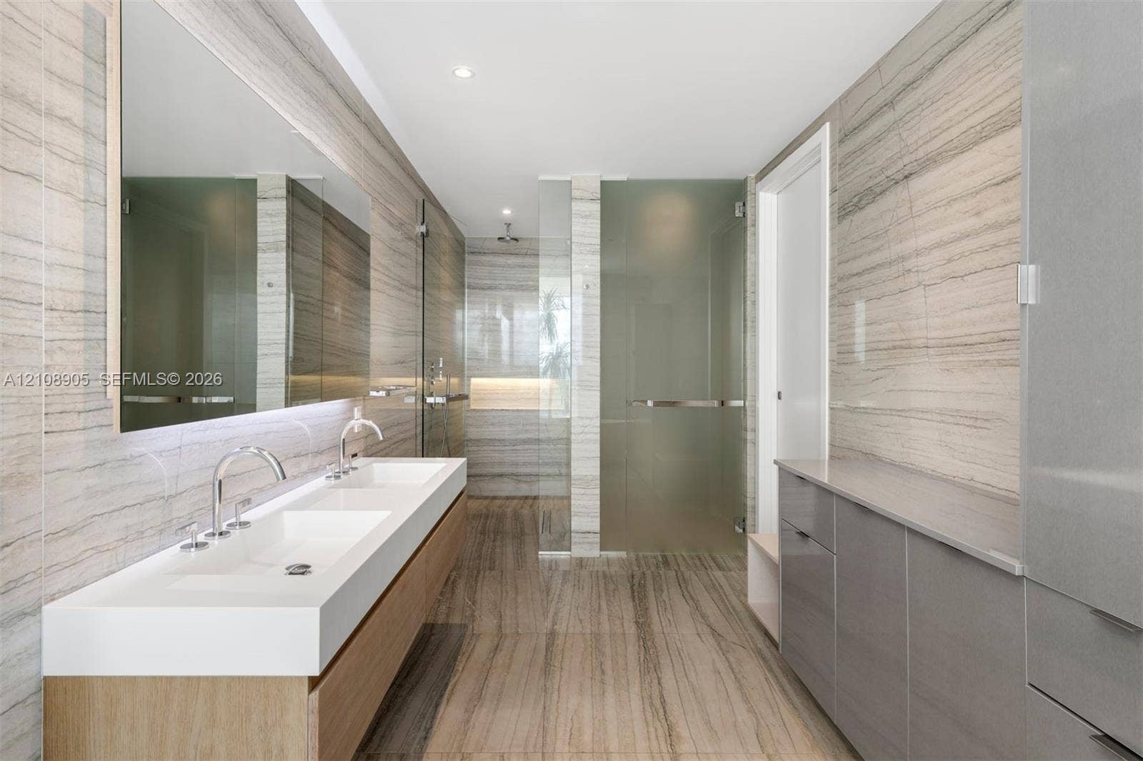Full-wall stone tiling, dual-sink floating vanity with polished chrome fixtures, oversized wall-mounted mirror, glass-enclosed shower, and floor-to-ceiling cabinetry