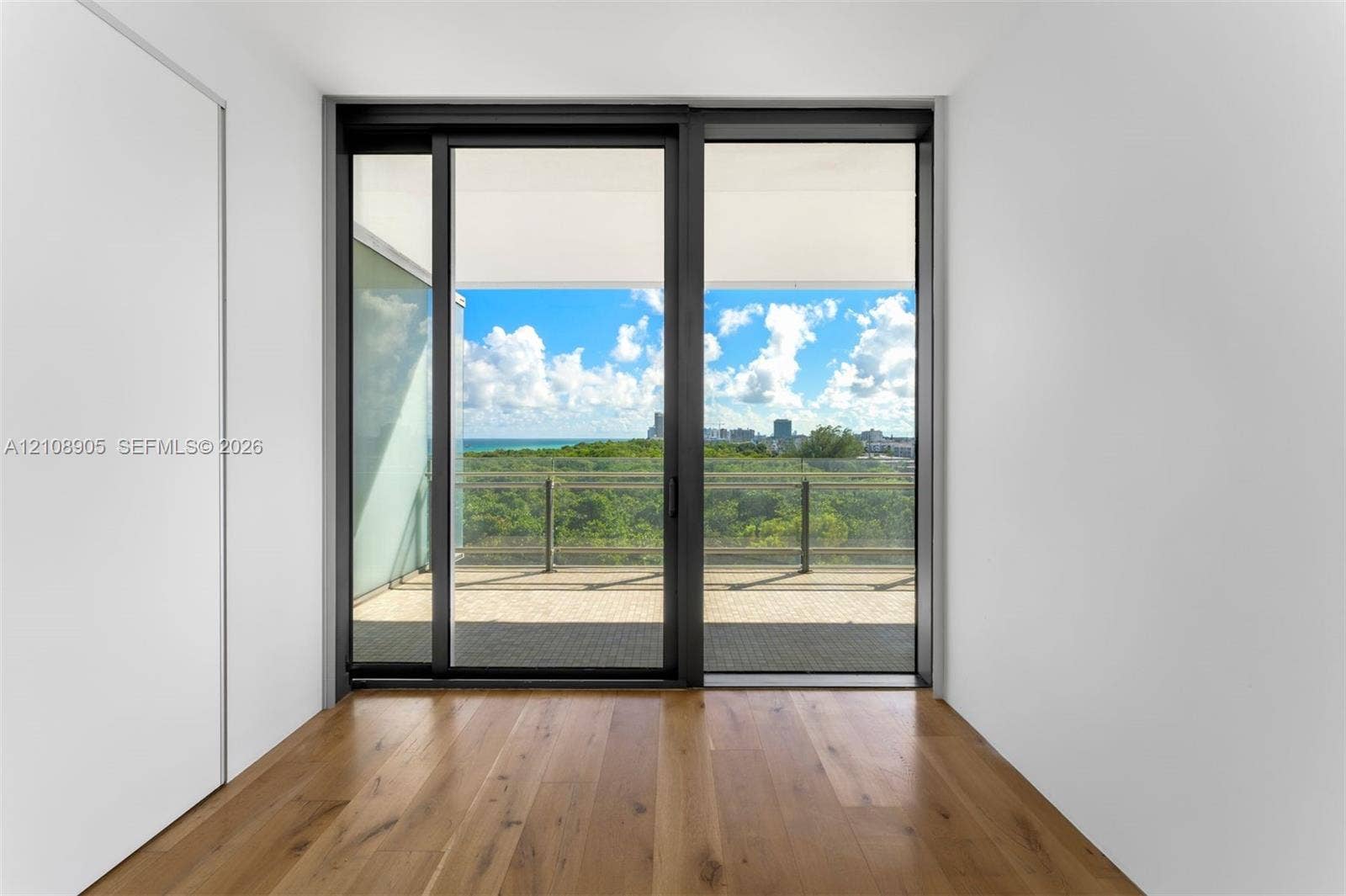 Floor-to-ceiling sliding glass doors, wood-finish flooring, private balcony with metal railing, and panoramic coastal and urban scenery