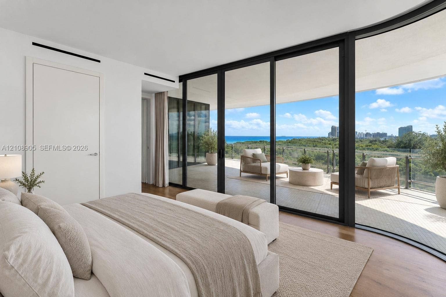 Floor-to-ceiling retractable glass walls, wide-plank wood flooring, expansive private balcony, recessed linear lighting, and coastal landscape backdrop
