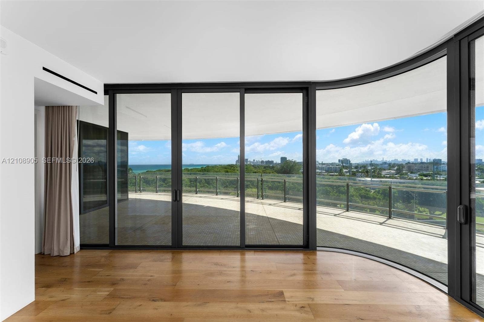 Curved floor-to-ceiling glass walls, expansive wraparound terrace, dark-framed sliding glass doors, and wide-plank wood-finish flooring