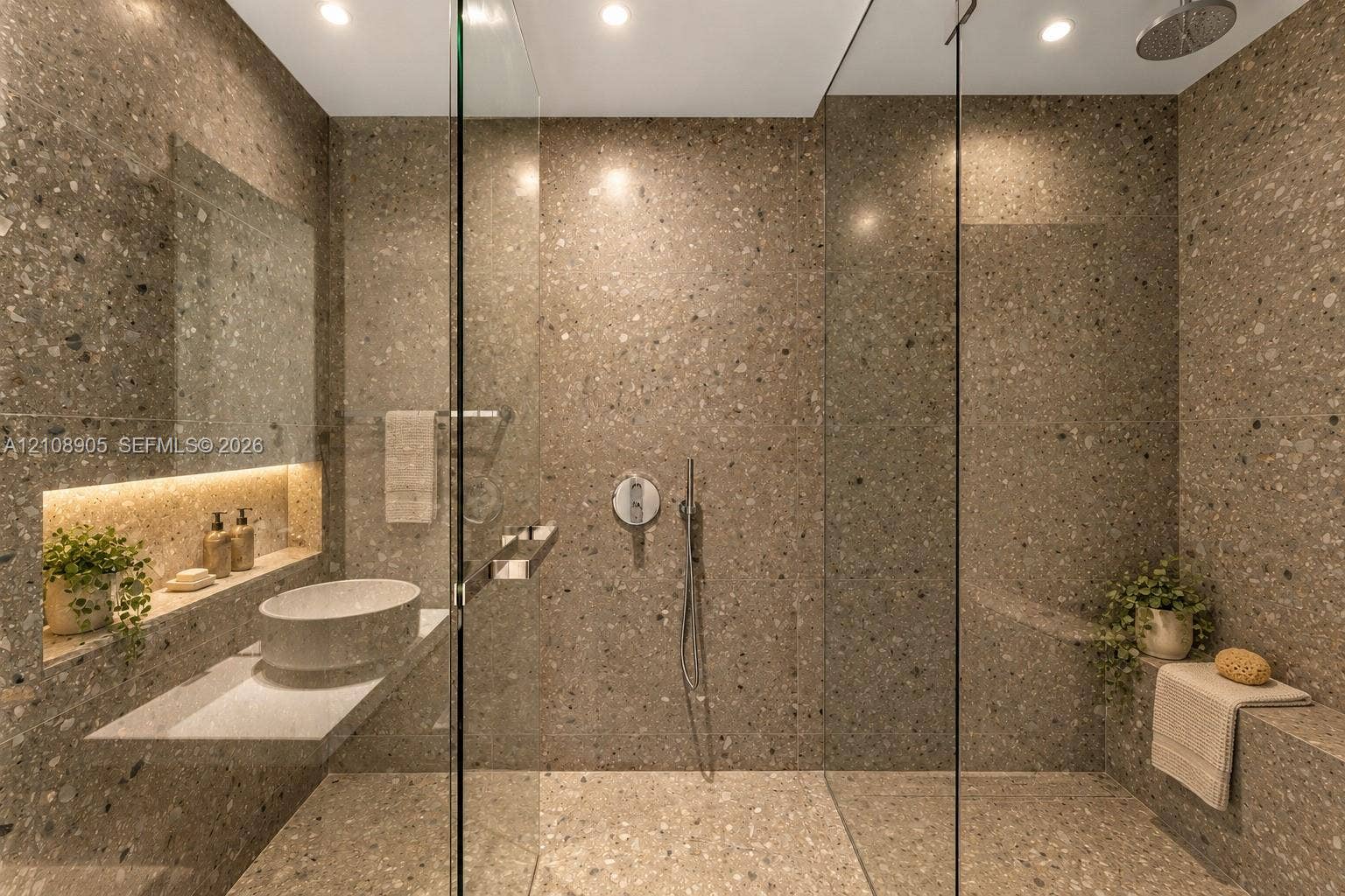 Full-height natural stone tiling, walk-in shower with frameless glass enclosure, wall-mounted faucet, floating vanity with vessel sink, and recessed integrated shelving with accent lighting