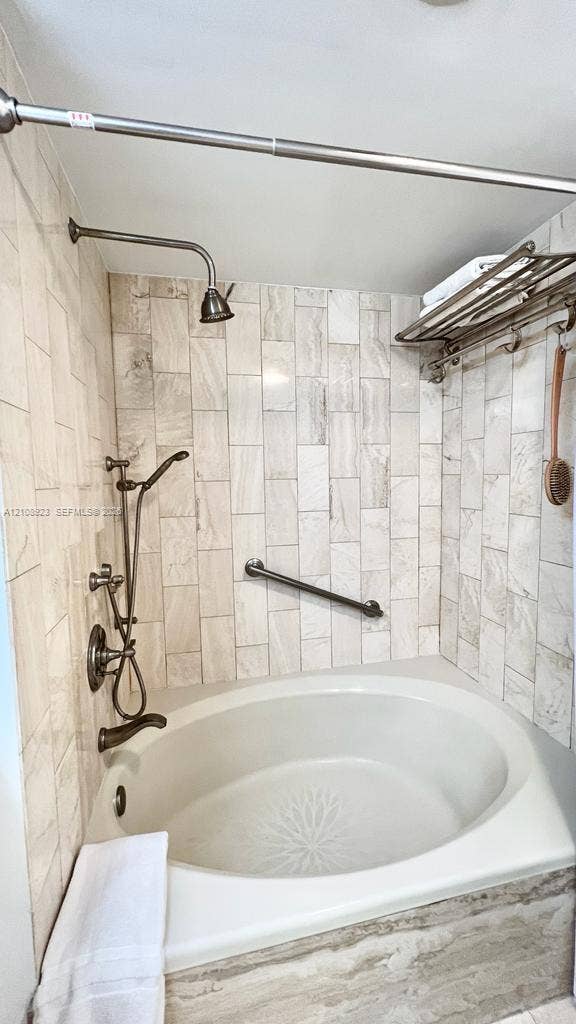 Oval soaking tub featuring a metal finish handheld shower head and rainfall fixture, offset by floor-to-ceiling neutral tile surround, a wall-mounted grab bar, and an integrated metal storage rack
