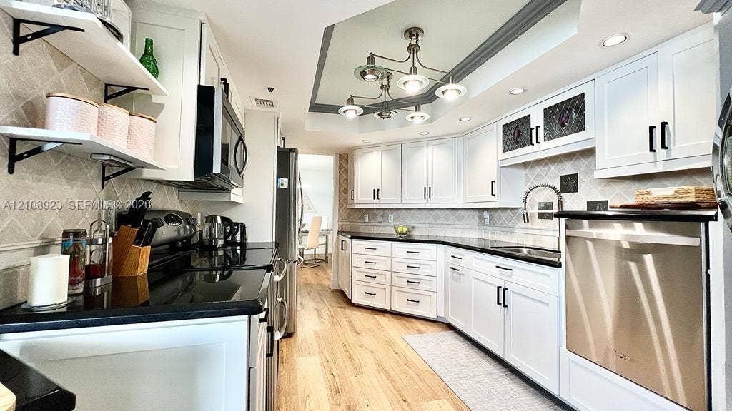 Kitchen featuring white shaker-style cabinetry, black countertops, wood-finish flooring, a multi-arm chandelier, and stainless steel appliances