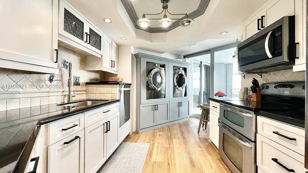 Kitchen featuring wood-finish flooring, white cabinetry with matte black hardware, stone-finish countertops, tile backsplash, and a multi-light tray ceiling fixture