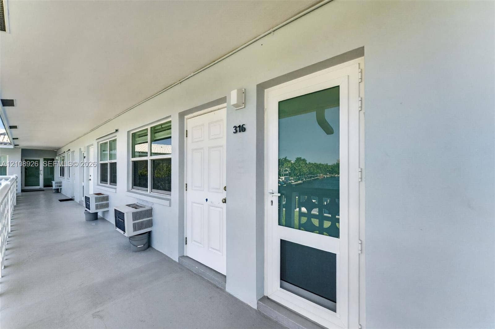 Covered exterior walkway featuring a six-panel entry door, glass-paneled storm door, dual-pane windows, and exterior-mounted cooling units