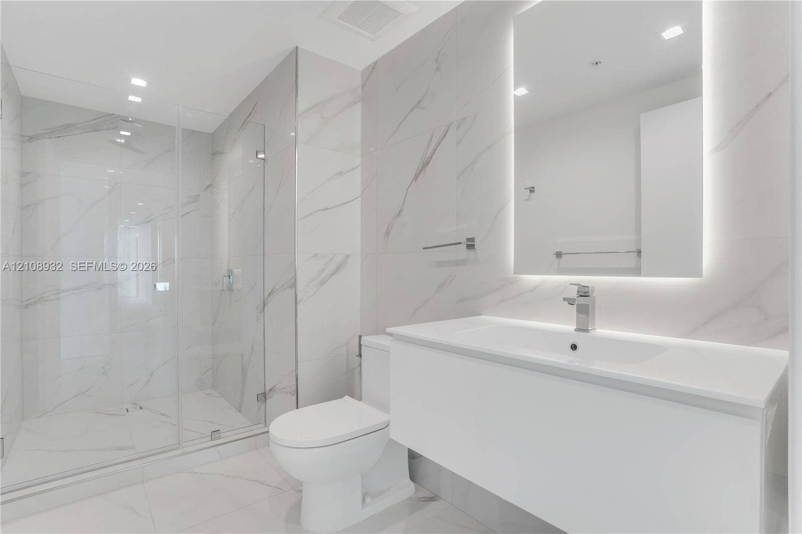 1170 E Hallandale Beach Boulevard, #1201, Hallandale Beach, FL 33009 Modern bathroom featuring floor-to-ceiling large-format stone-patterned tile, a glass-enclosed walk-in shower, a white floating vanity with a seamless countertop, and a wall-mounted mirror with integrated backlighting